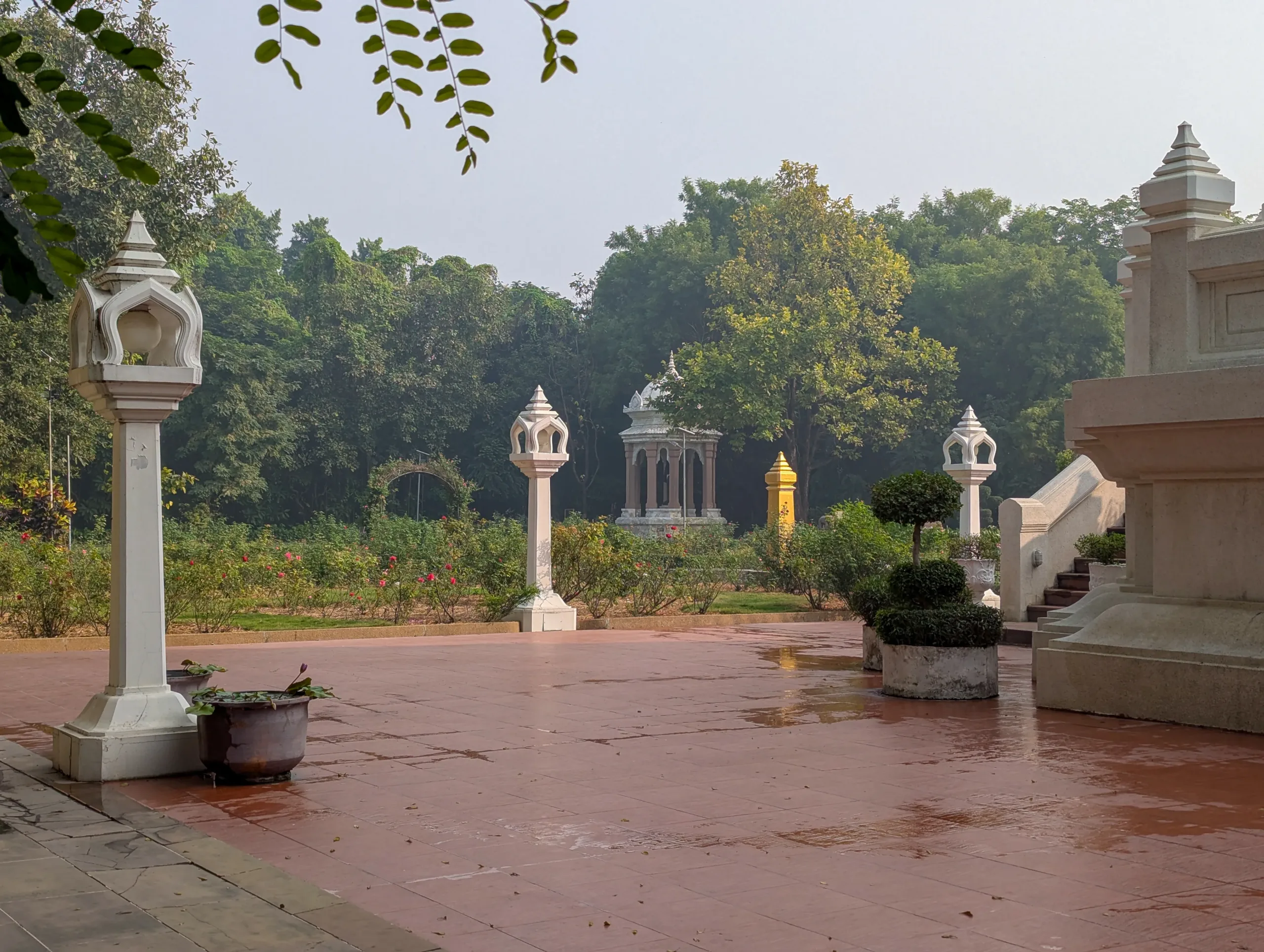 Close view of Wat Thai Temple’s garden in Kushinagar with white ornamental lantern posts, manicured topiary, lotus pots, rose beds, and a small pavilion amid lush trees.
