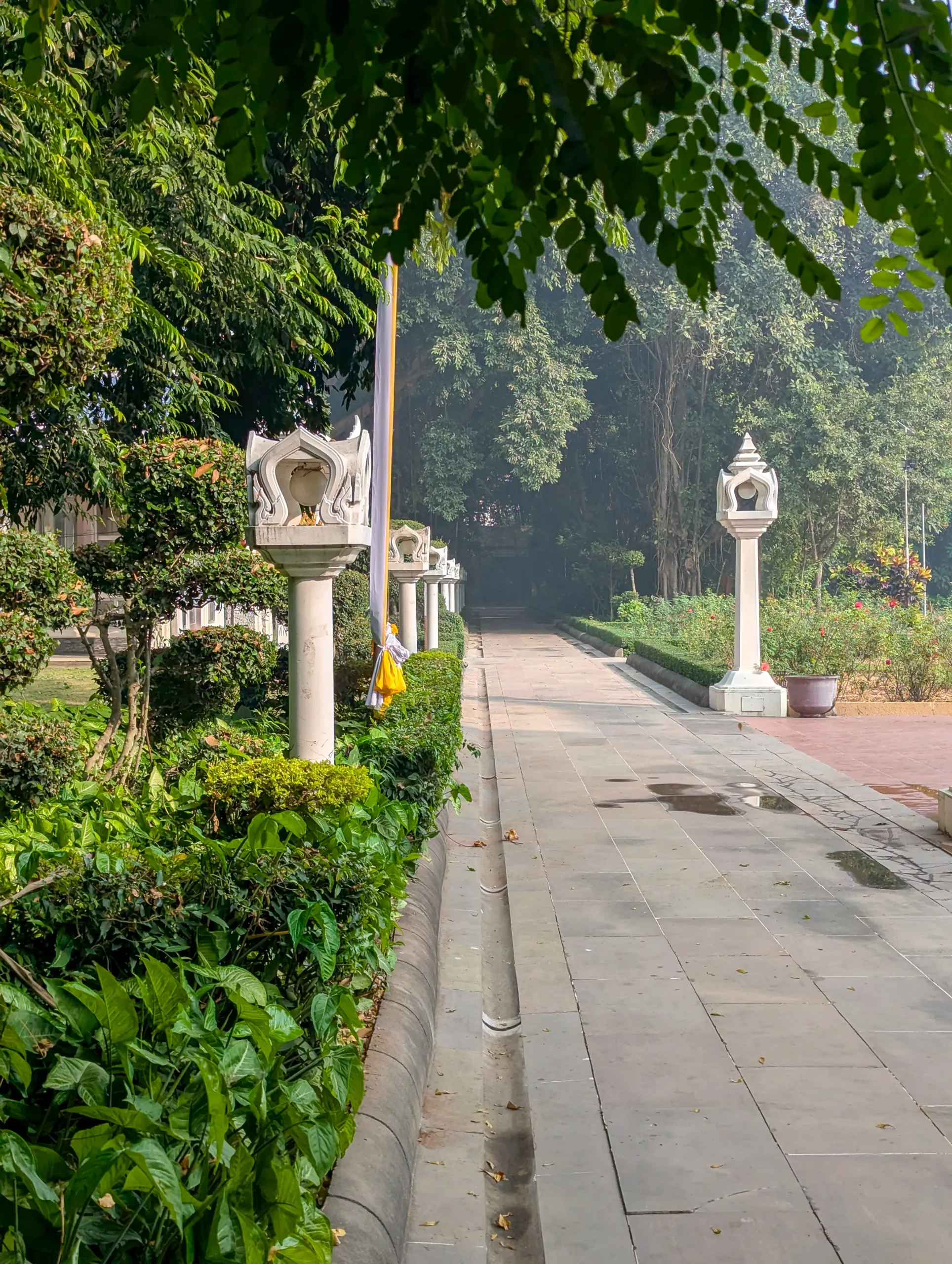 Stone pathway at Wat Thai Temple in Kushinagar bordered by lush hedges and white ornamental lantern posts under overhanging tree branches.