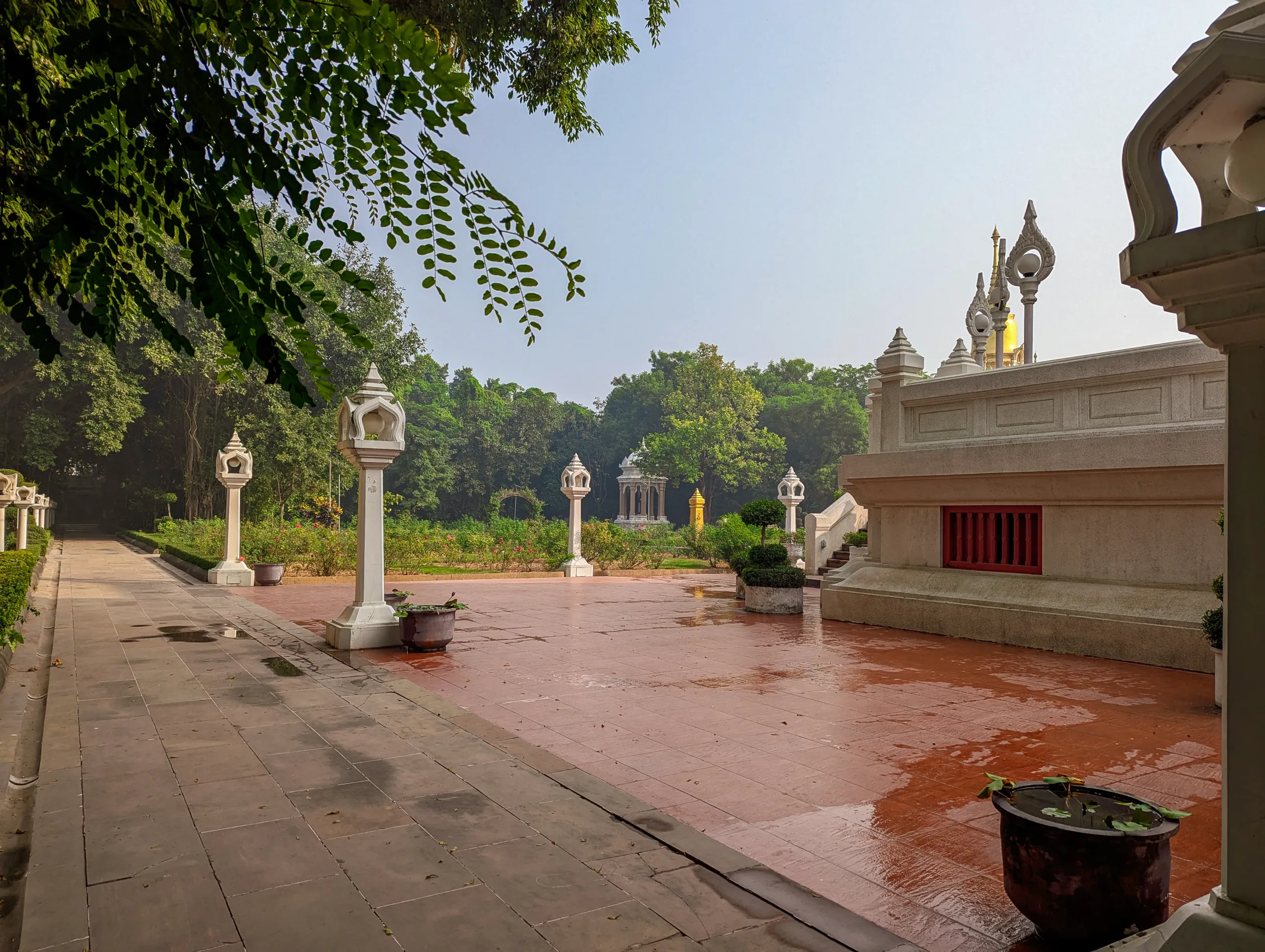 Peaceful garden courtyard at Wat Thai Temple in Kushinagar with white ornamental lamp posts, trimmed hedges, potted plants, and a temple structure with red windows.