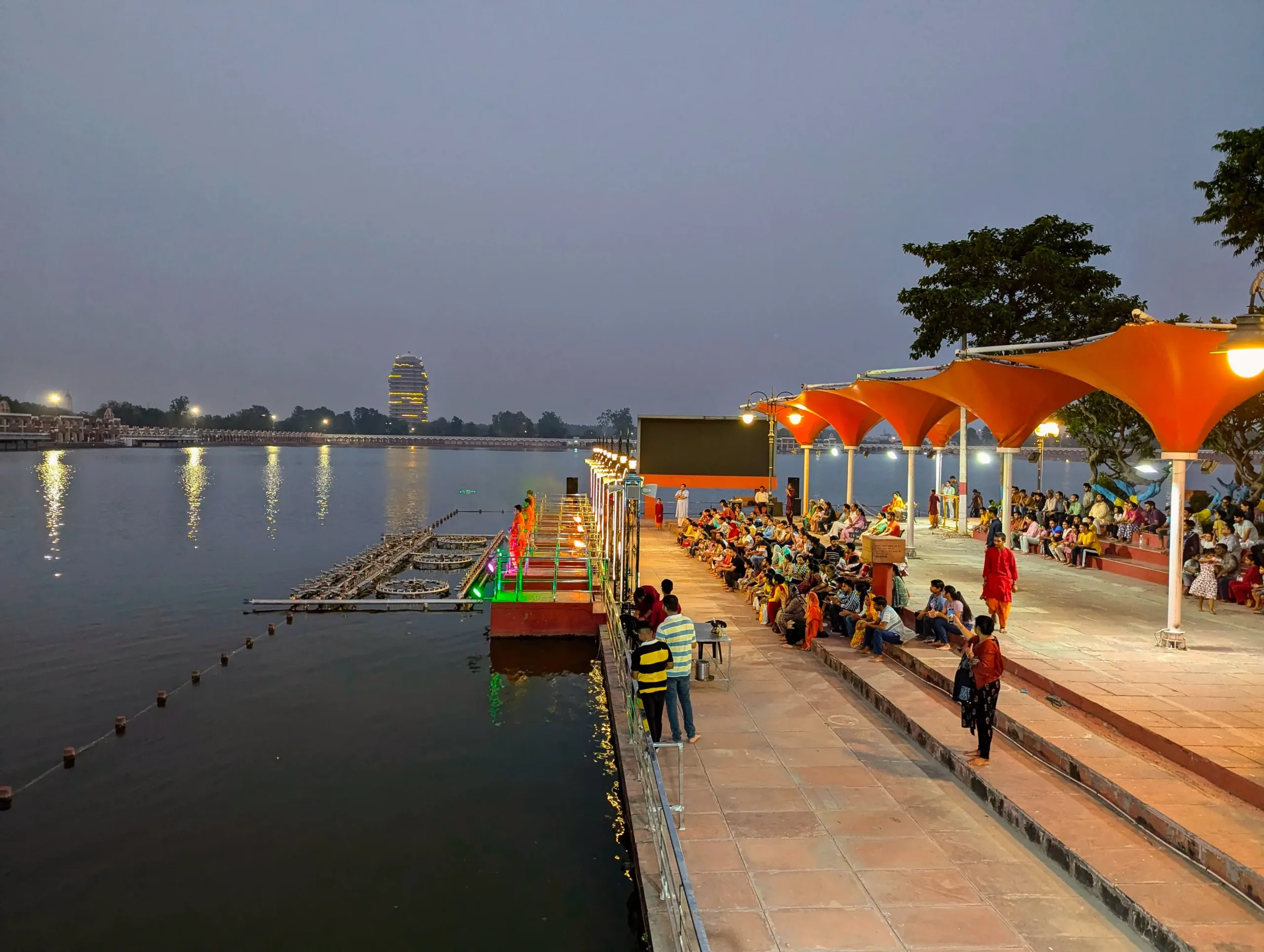 Crowds seated under orange canopies on the Brahma Sarovar ghats in Kurukshetra with colorful lights on the aarti platform at dusk