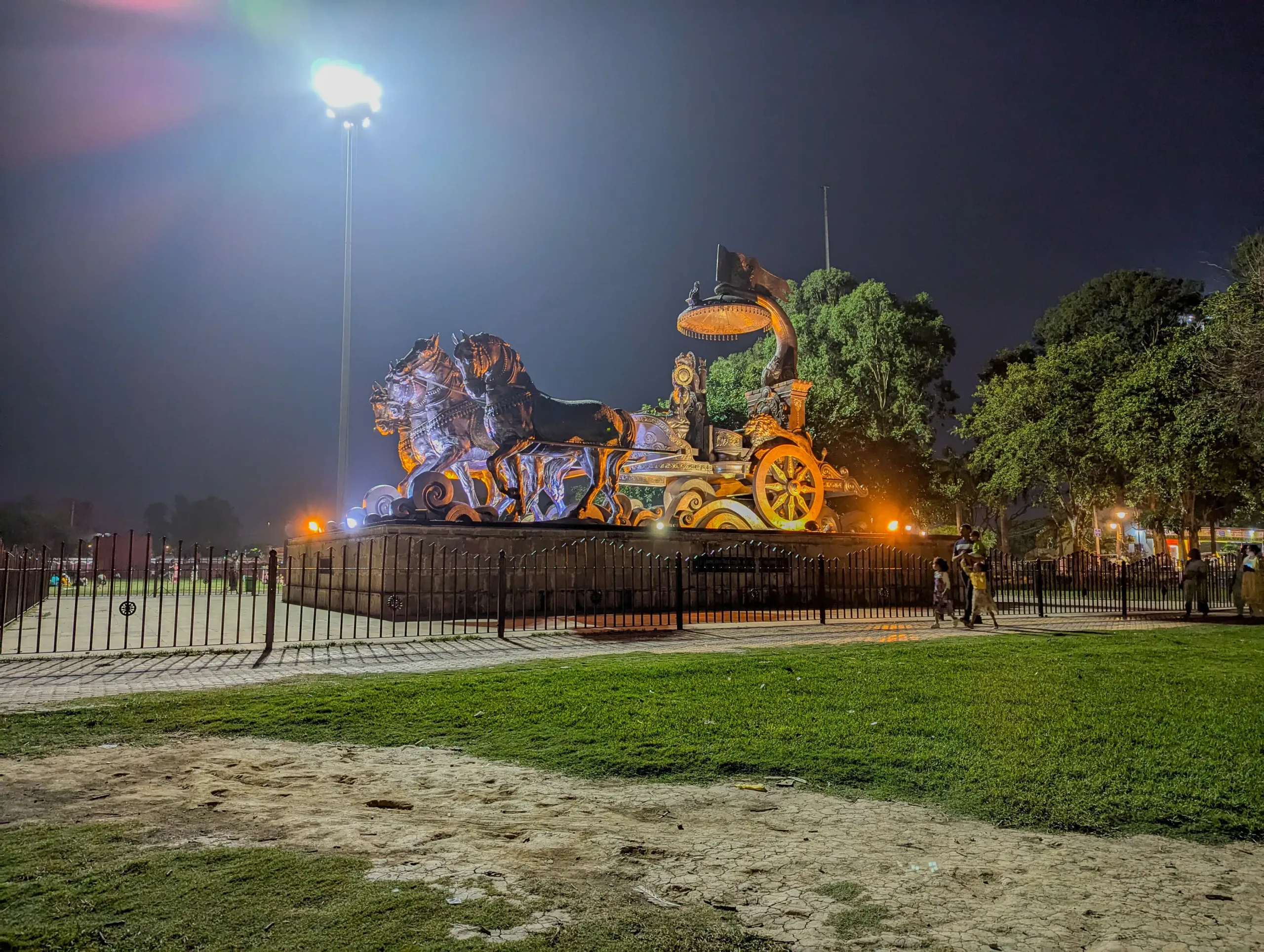 Close night view of the illuminated Krishna and Arjuna chariot statue at Brahma Sarovar in Kurukshetra, fenced lawn in foreground