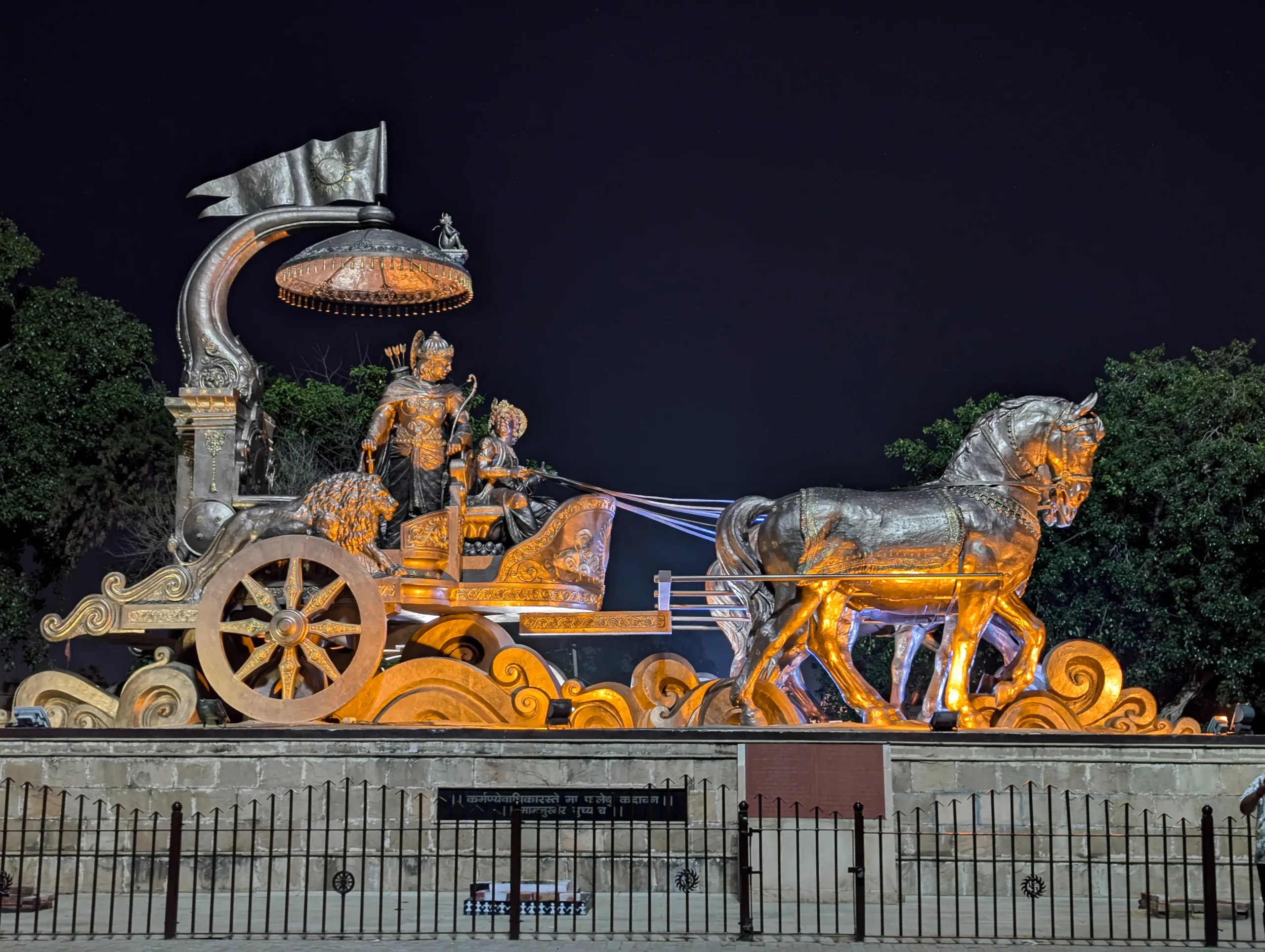 Front night view of the illuminated bronze chariot statue of Krishna and Arjuna with horses at Brahma Sarovar in Kurukshetra, seen behind a fence