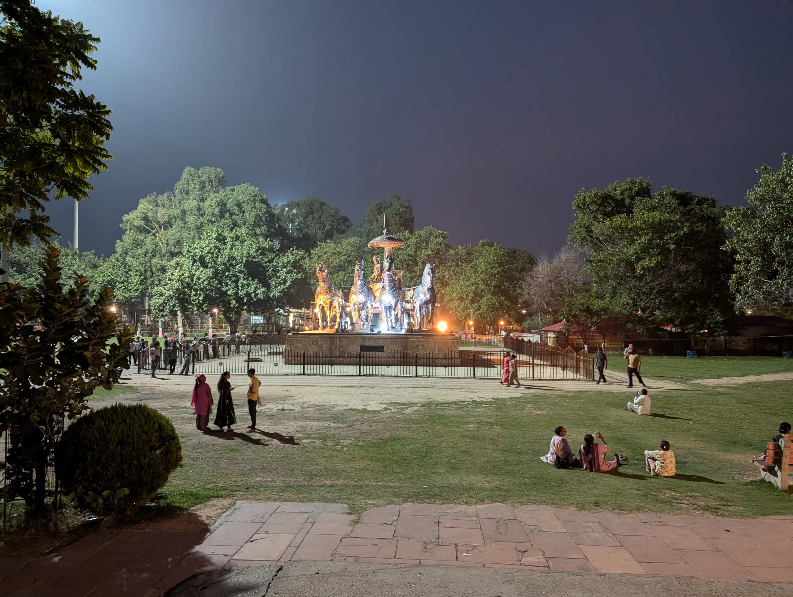 Illuminated Krishna and Arjuna chariot statue at Brahma Sarovar in Kurukshetra with people relaxing on the lawn at night