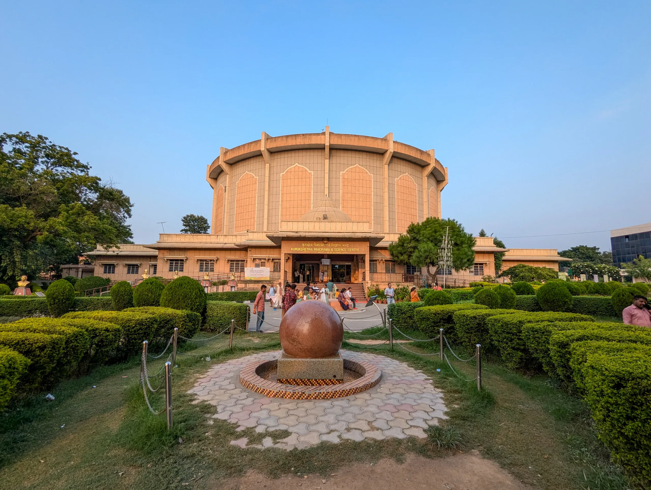 Front view of the cylindrical Kurukshetra Panorama & Science Centre with visitors and a floating granite ball fountain in the garden