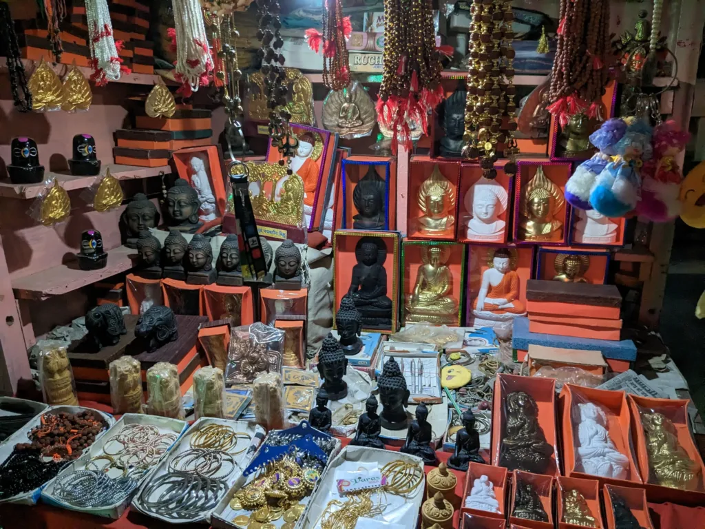 Nighttime view of a Sarnath shop in Banaras displaying Buddha busts and statues, prayer beads, metal pendants, and handmade souvenirs arranged in bright boxes and trays