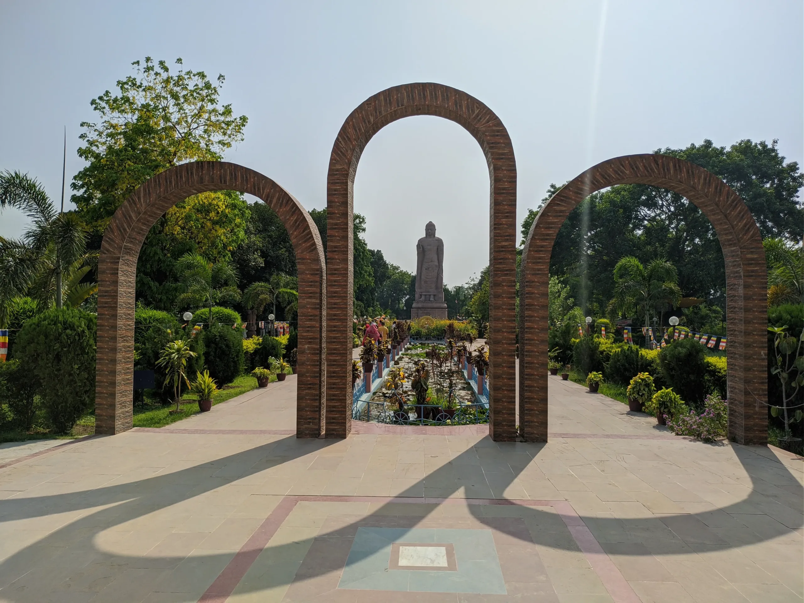 Symmetrical view at Sarnath’s Mulagandha Kuti Vihar in Banaras showing three brick arches framing a landscaped garden and a standing Buddha statue beneath a bright sky