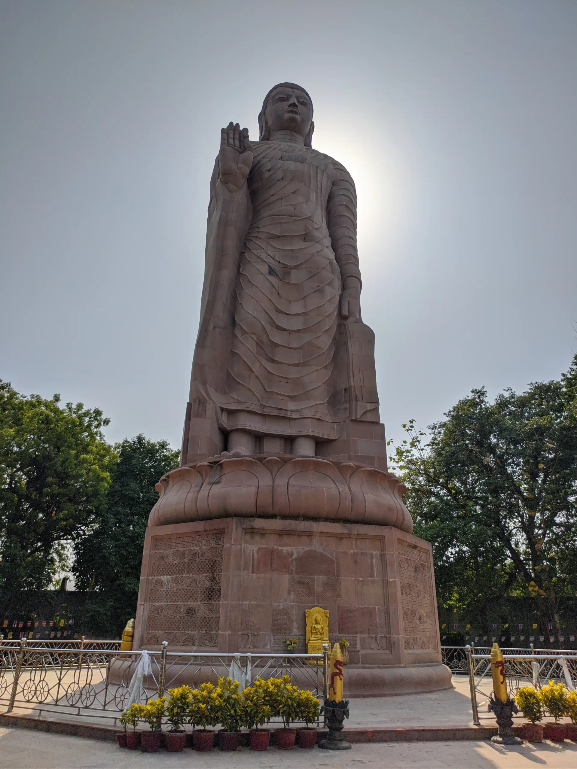 Low-angle portrait of the tall standing Buddha statue at Sarnath’s Mulagandha Kuti Vihar near Banaras, backlit so the sun forms a soft halo behind the head, with garden trees around